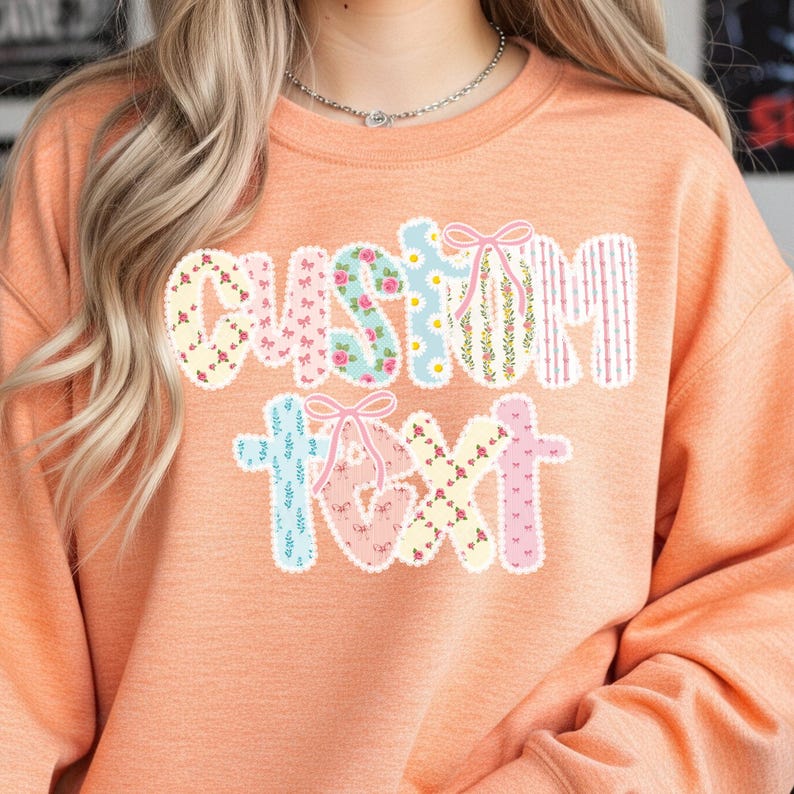 May include: Peach-colored sweatshirt with the words "CUSTOM TEXT" in a patchwork design. The letters are decorated with floral patterns, bows, and stripes. The sweatshirt is a casual, comfortable style.