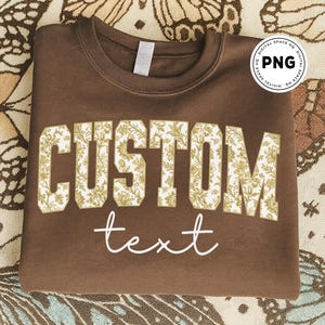 May include: A brown sweatshirt with the word "CUSTOM" in large, floral-patterned letters and the word "text" in a cursive font. The floral pattern features gold and white flowers. The sweatshirt is folded on a patterned surface.