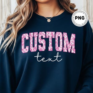 May include: Navy blue sweatshirt with the word "CUSTOM" in large, pink floral print letters. Below, the word "text" is written in a cursive font. The sweatshirt is a solid color.