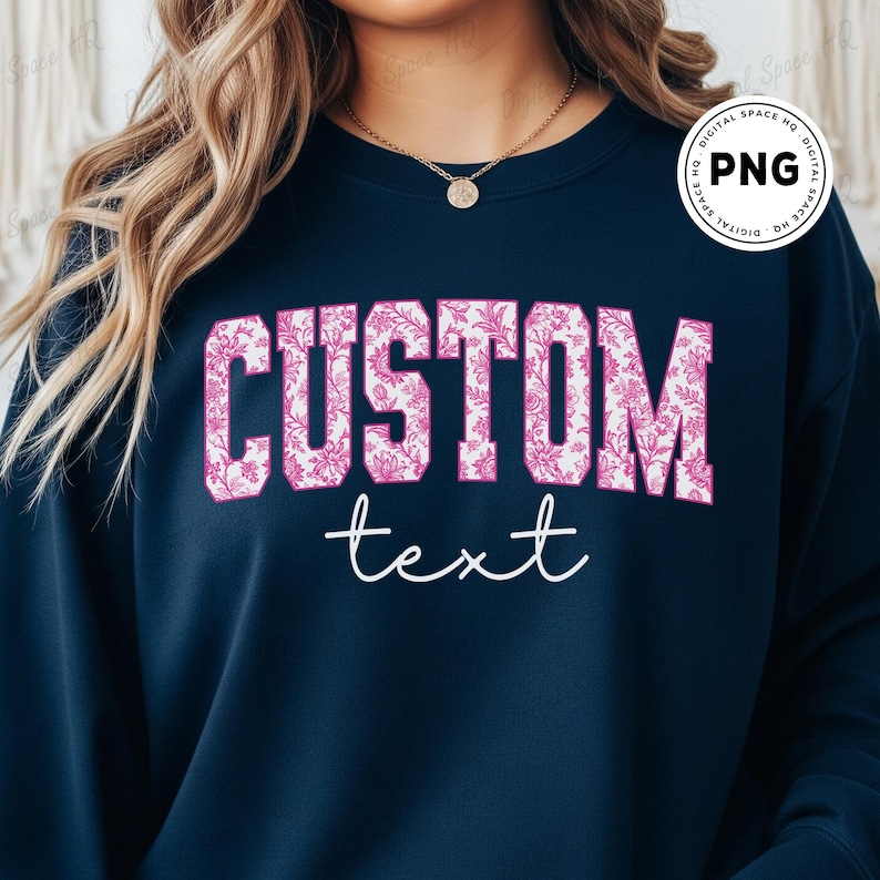 May include: Navy blue sweatshirt with the word "CUSTOM" in large, pink floral print letters. Below, the word "text" is written in a cursive font. The sweatshirt is a solid color.