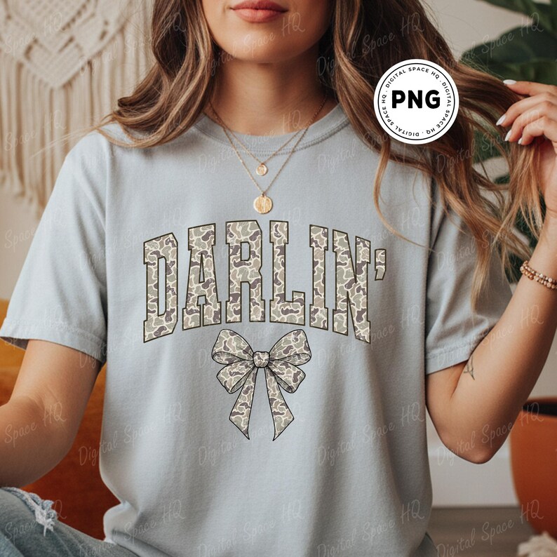 May include: A light blue t-shirt with the word "DARLIN'" in a camouflage pattern across the chest. Below the text is a bow with a matching camouflage design. The shirt is a casual style.