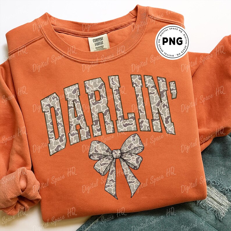 May include: Orange sweatshirt with the word "DARLIN'" in a camouflage pattern. A bow graphic is below the text. The sweatshirt has a crew neck and long sleeves.