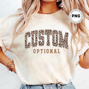 May include: A cream-colored sweatshirt with the words "CUSTOM" in a leopard print design and the word "OPTIONAL" in a brown font. The sweatshirt is a casual style, suitable for everyday wear.