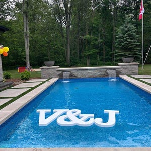 Peut inclure: Un grand panneau blanc flottant affichant les lettres V&J dans une piscine rectangulaire à l'eau bleue. La piscine dispose d'un mur en pierre avec deux fontaines intégrées, entouré d'une terrasse en béton et d'une forêt verte en arrière-plan.