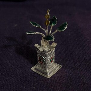 Puede incluir: Una escultura decorativa plateada de una planta. La escultura presenta una base cuadrada y un jarrón detallado y estampado que sostiene una planta con hojas verdes y detalles amarillos. La escultura mide aproximadamente 7,6 cm de altura.