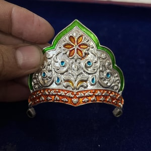 Peut inclure: Couronne argentée ornée avec des détails floraux et des volutes. Elle présente des bordures et des accents en émail vert, orange et jaune, ainsi que de petites pierres bleues. La couronne a deux petits pieds.