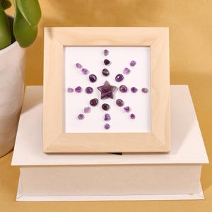 May include: A square light-colored wooden frame containing a white background with a star-shaped purple amethyst stone at the center, surrounded by a radial pattern of smaller, tumbled purple amethyst stones arranged in a starburst design.