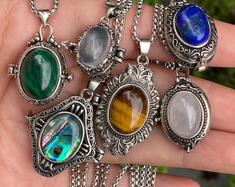 Puede incluir: Una colección de colgantes de metal plateado en cadenas, cada uno con una piedra preciosa ovalada diferente, incluyendo malaquita verde, abulón iridiscente, ojo de tigre dorado, lapislázuli azul y cuarzo translúcido.