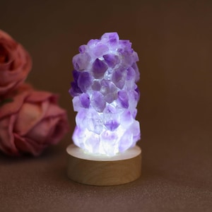 May include: A cylindrical lamp composed of numerous raw, translucent purple amethyst crystal points clustered together. The crystals are illuminated from within by a bright white light and are mounted on a smooth, circular light-coloured wooden base.