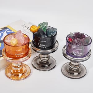 Op de afbeelding: Drie glazen oliediffusers op voetstukken, gevuld met ruwe kristallen en gedroogde bloemen. De amberkleurige, zwarte en paarse glazen houders zijn voorzien van de witte tekst OIL DIFFUSER CRYSTAL AROMA. Elk staat op een bijpassende glazen standaard.