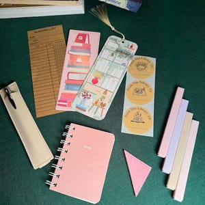May include: A collection of stationery items including a pink spiral notebook with the word "NOTES", bookmarks, stickers, and highlighters. A copy of "Pride and Prejudice" is visible in the background. The items are arranged on a green surface.
