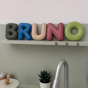 Op de afbeelding: Een set van vijf individuele, afgeronde, glanzende letters die de naam BRUNO vormen in donkergroen, blauw, beige, roze en lichtgroen, geplaatst op een plank boven een speelgoedkeuken met een zilveren kraan, een kleine plant en een kookpot.