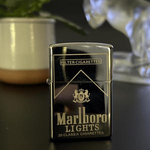 Puede incluir: Un encendedor de metal negro con las palabras "Marlboro Lights" y "Filter Cigarettes" impresas. El encendedor tiene un acabado brillante y una forma rectangular cl&aacute;sica. El dise&ntilde;o incluye un logotipo con dos leones y el texto "20 CLASS A CIGARETTES".