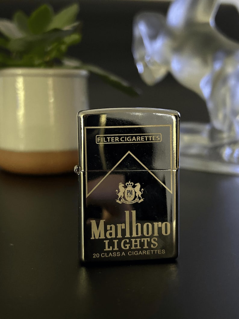 Puede incluir: Un encendedor de metal negro con las palabras "Marlboro Lights" y "Filter Cigarettes" impresas. El encendedor tiene un acabado brillante y una forma rectangular cl&aacute;sica. El dise&ntilde;o incluye un logotipo con dos leones y el texto "20 CLASS A CIGARETTES".