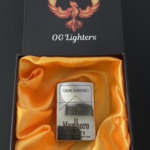 Puede incluir: Un encendedor plateado Marlboro Lights en una caja negra abierta con forro de sat&eacute;n naranja. La caja tiene el texto "OG Lighters" y un logotipo de f&eacute;nix. El encendedor tiene las palabras "FILTER CIGARETTES" y "Marlboro LIGHTS".