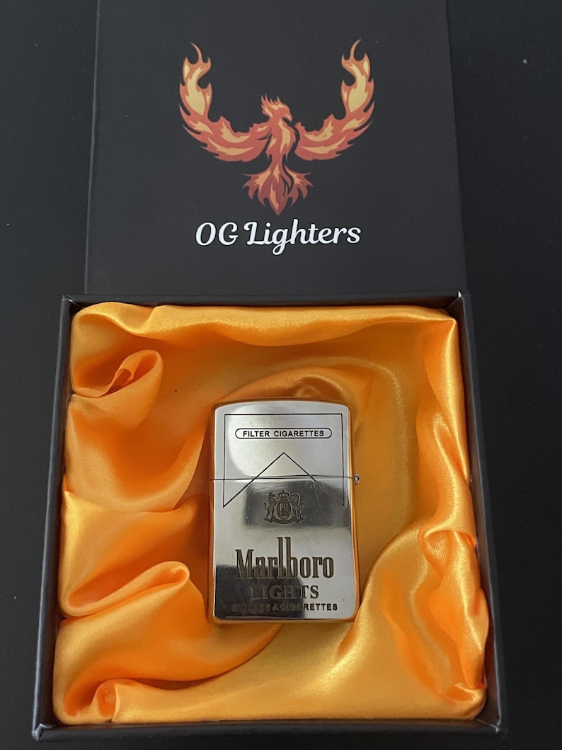 Puede incluir: Un encendedor plateado Marlboro Lights en una caja negra abierta con forro de sat&eacute;n naranja. La caja tiene el texto "OG Lighters" y un logotipo de f&eacute;nix. El encendedor tiene las palabras "FILTER CIGARETTES" y "Marlboro LIGHTS".