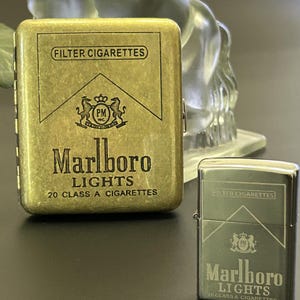 Puede incluir: Dos pitilleras met&aacute;licas, una rectangular y otra con forma de encendedor, ambas con el logotipo y el texto "Marlboro Lights". La pitillera rectangular es dorada, mientras que el encendedor es plateado. Ambas tienen el texto "FILTER CIGARETTES".