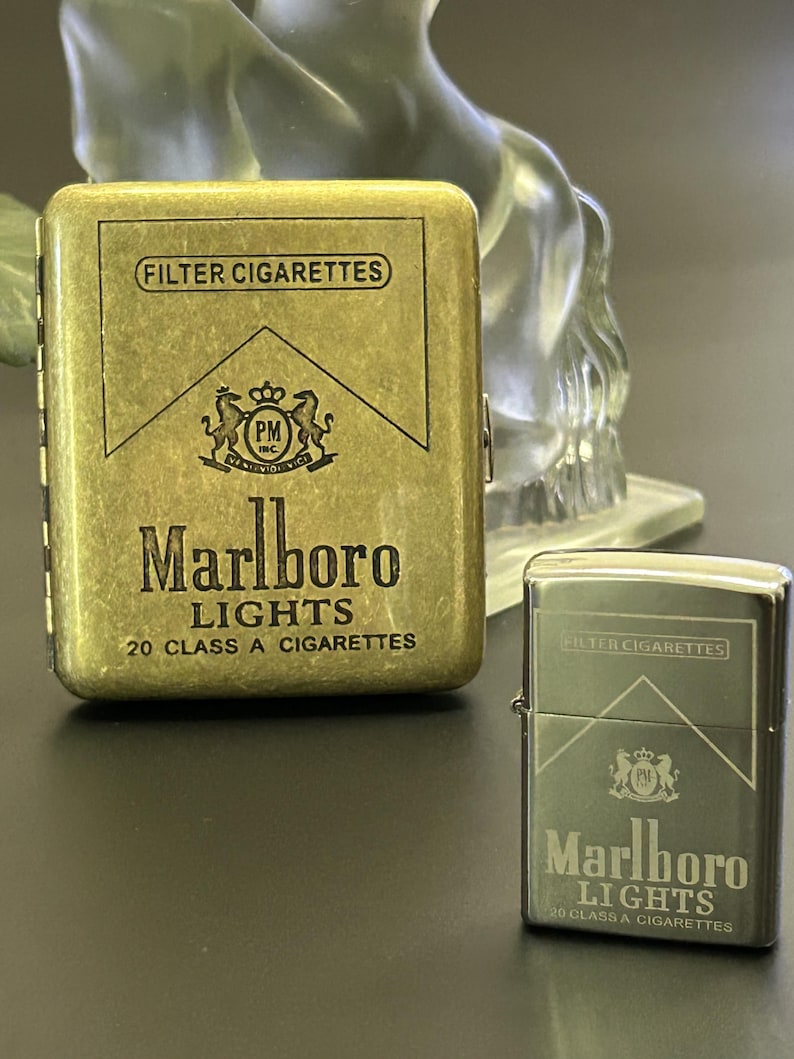 Puede incluir: Dos pitilleras met&aacute;licas, una rectangular y otra con forma de encendedor, ambas con el logotipo y el texto "Marlboro Lights". La pitillera rectangular es dorada, mientras que el encendedor es plateado. Ambas tienen el texto "FILTER CIGARETTES".