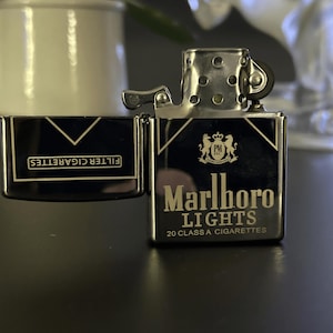 Puede incluir: Un encendedor Marlboro Lights negro y plateado con la tapa abierta, que muestra el nombre de la marca y el texto "20 CLASS A CIGARETTES". El encendedor est&aacute; sobre una superficie oscura. Las palabras "FILTER CIGARETTES" son visibles en la tapa.