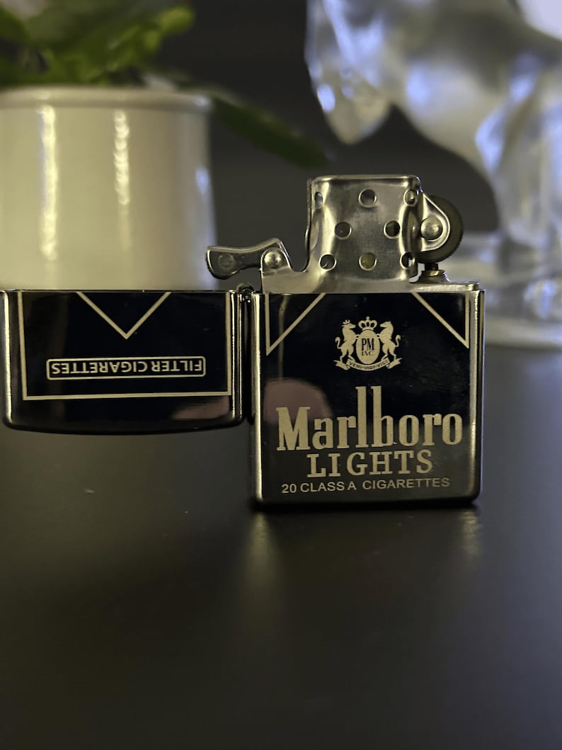 Puede incluir: Un encendedor Marlboro Lights negro y plateado con la tapa abierta, que muestra el nombre de la marca y el texto "20 CLASS A CIGARETTES". El encendedor est&aacute; sobre una superficie oscura. Las palabras "FILTER CIGARETTES" son visibles en la tapa.