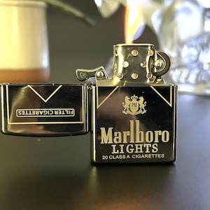 Puede incluir: Un encendedor de metal negro y dorado con las palabras "Marlboro Lights" y "20 Class A Cigarettes" impresas. El encendedor est&aacute; abierto y encendido, con una llama visible. Un estuche de cigarrillos a juego est&aacute; al lado del encendedor.