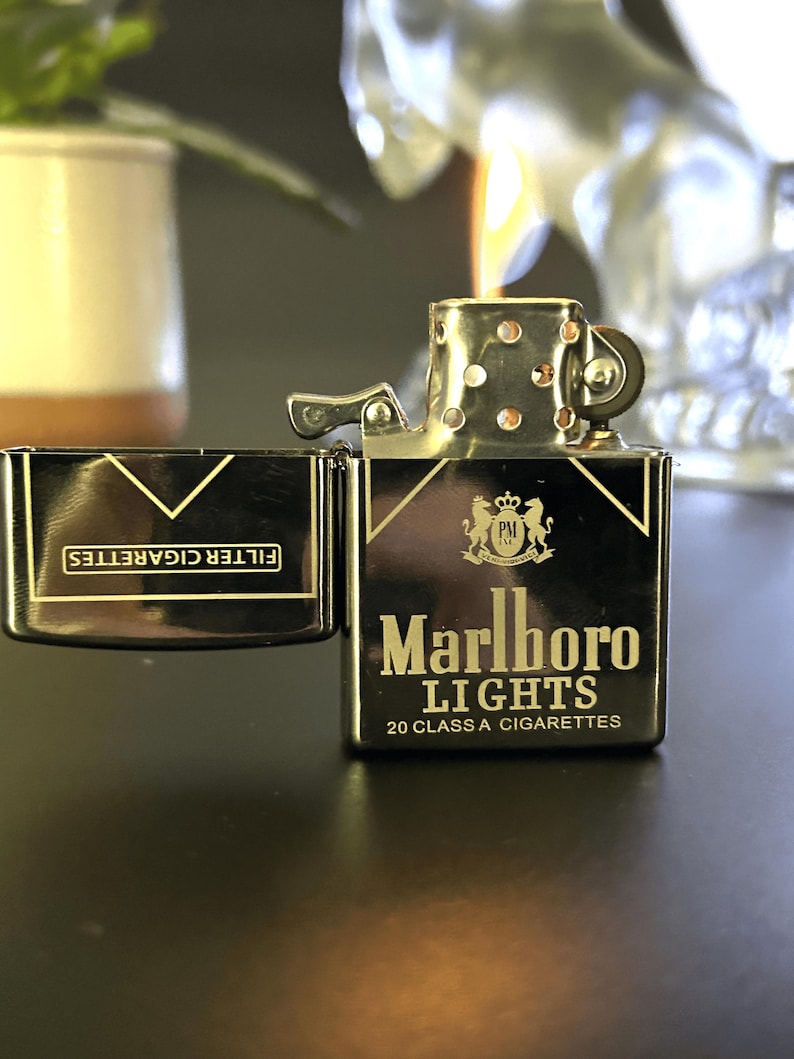Puede incluir: Un encendedor de metal negro y dorado con las palabras "Marlboro Lights" y "20 Class A Cigarettes" impresas. El encendedor est&aacute; abierto y encendido, con una llama visible. Un estuche de cigarrillos a juego est&aacute; al lado del encendedor.