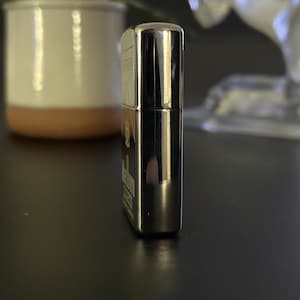 Puede incluir: Un encendedor cromado con el logotipo de Marlboro Lights. El encendedor rectangular tiene bordes redondeados y una superficie brillante y reflectante. El encendedor est&aacute; sobre una superficie oscura.