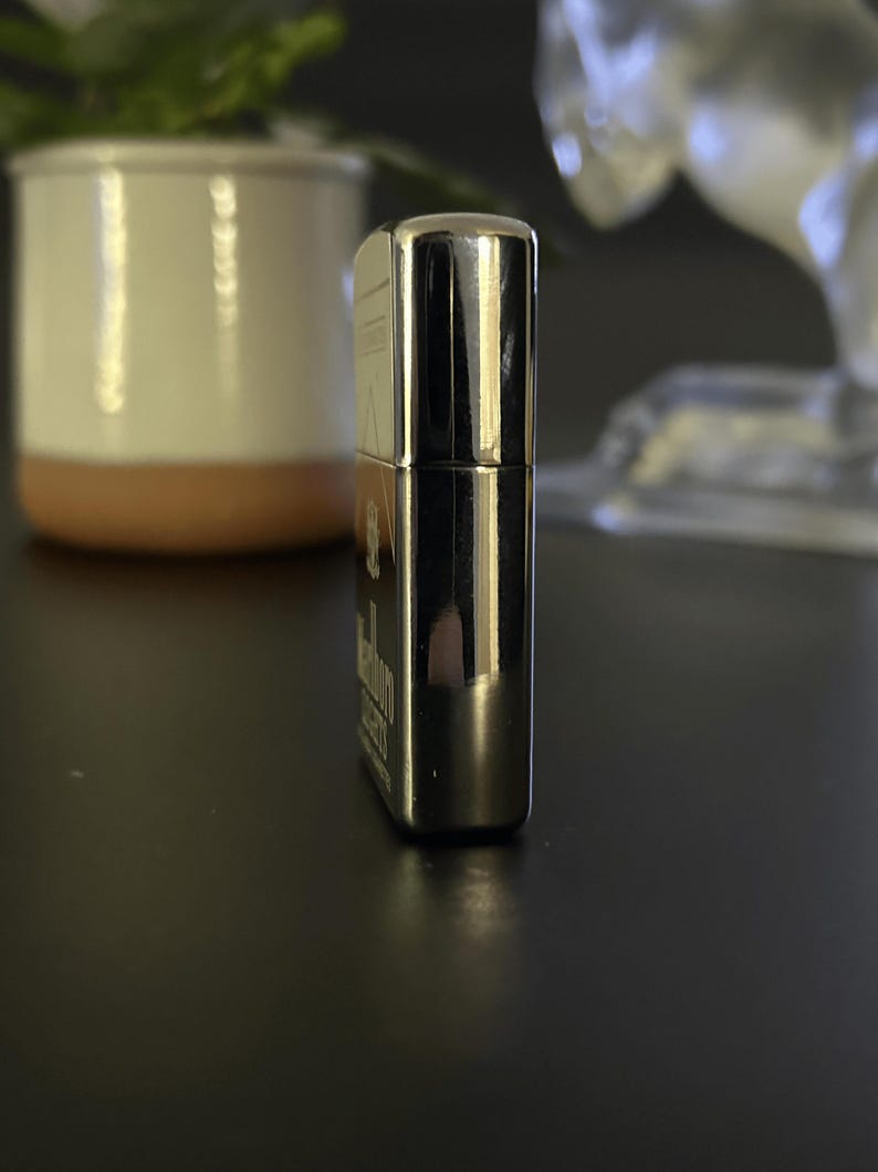 Puede incluir: Un encendedor cromado con el logotipo de Marlboro Lights. El encendedor rectangular tiene bordes redondeados y una superficie brillante y reflectante. El encendedor est&aacute; sobre una superficie oscura.