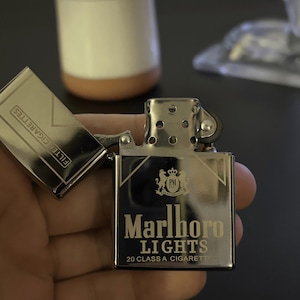 Puede incluir: Un encendedor Marlboro Lights plateado con la tapa abierta, mostrando "FILTER CIGARETTES". El encendedor presenta el logotipo de Marlboro Lights y el texto "20 CLASS A CIGARETTES."