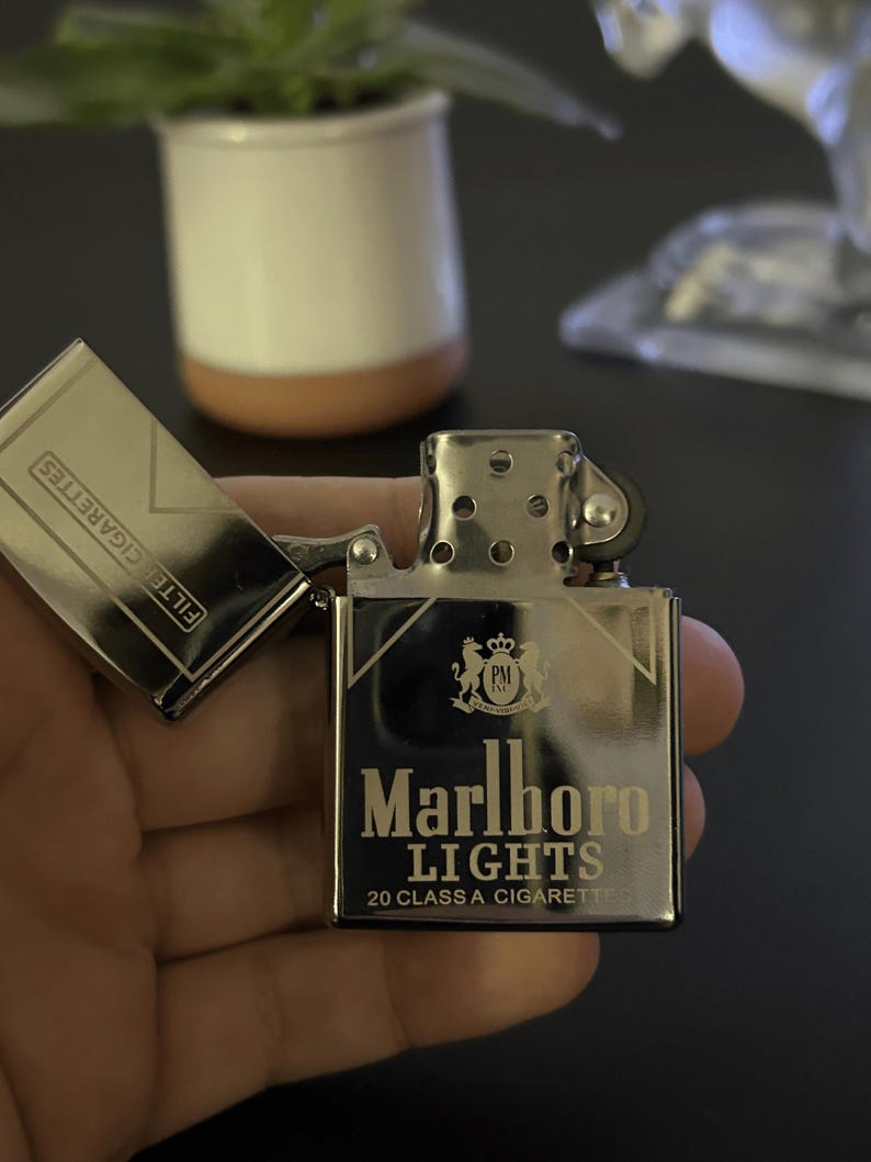 Puede incluir: Un encendedor Marlboro Lights plateado con la tapa abierta, mostrando "FILTER CIGARETTES". El encendedor presenta el logotipo de Marlboro Lights y el texto "20 CLASS A CIGARETTES."