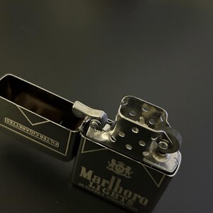 Puede incluir: Un encendedor de metal negro con el logotipo de Marlboro Lights en blanco. El encendedor est&aacute; abierto, revelando el compartimento para cigarrillos. El encendedor est&aacute; sobre una superficie oscura.