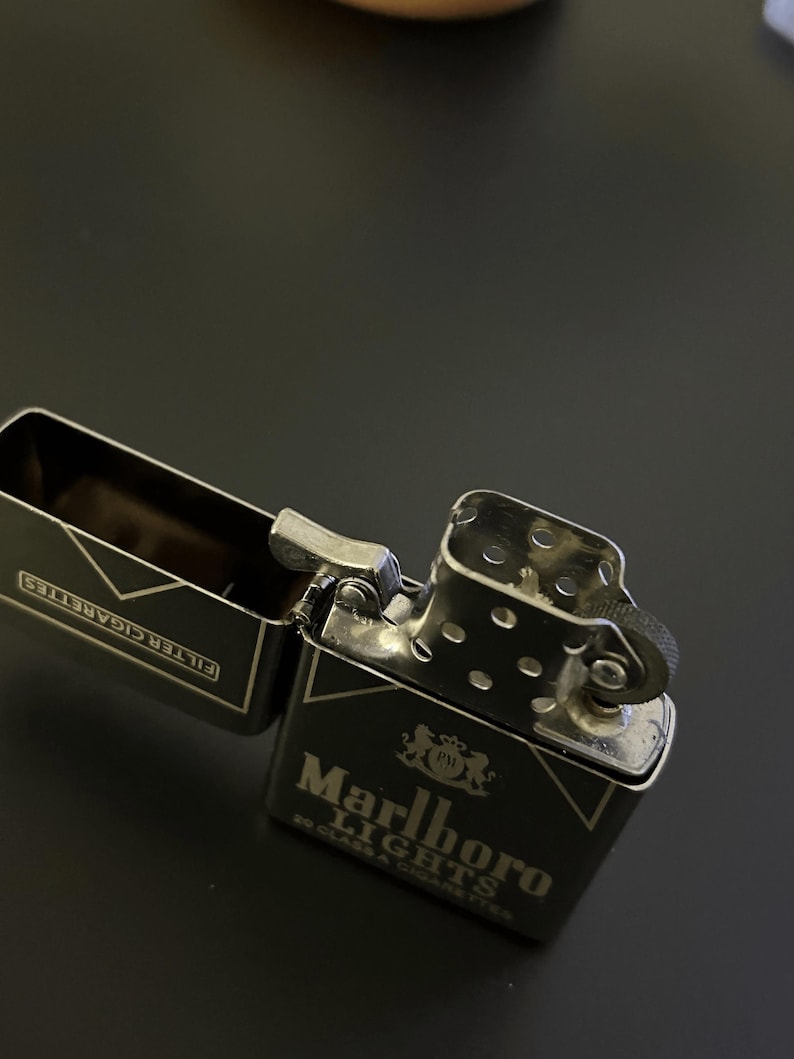 Puede incluir: Un encendedor de metal negro con el logotipo de Marlboro Lights en blanco. El encendedor est&aacute; abierto, revelando el compartimento para cigarrillos. El encendedor est&aacute; sobre una superficie oscura.
