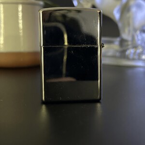 Puede incluir: Un encendedor plateado brillante de forma rectangular. El encendedor tiene una superficie lisa y reflectante y una peque&ntilde;a bisagra en el lateral. El fondo es oscuro, con un toque de planta y un objeto decorativo.