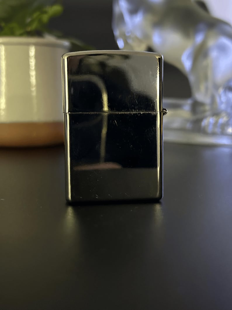 Puede incluir: Un encendedor plateado brillante de forma rectangular. El encendedor tiene una superficie lisa y reflectante y una peque&ntilde;a bisagra en el lateral. El fondo es oscuro, con un toque de planta y un objeto decorativo.