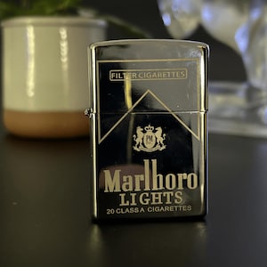 Puede incluir: Un encendedor cromado con el logotipo de Marlboro Lights. El encendedor tiene las palabras "FILTER CIGARETTES" en la parte superior y "Marlboro LIGHTS 20 CLASS A CIGARETTES" en la parte inferior. El fondo es negro.