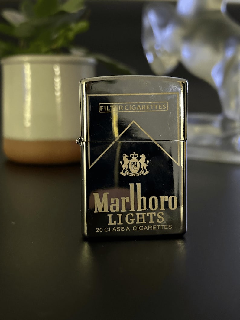 Puede incluir: Un encendedor cromado con el logotipo de Marlboro Lights. El encendedor tiene las palabras "FILTER CIGARETTES" en la parte superior y "Marlboro LIGHTS 20 CLASS A CIGARETTES" en la parte inferior. El fondo es negro.