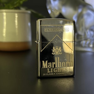 Puede incluir: Un encendedor plateado con el logotipo de Marlboro Lights. El encendedor tiene las palabras "FILTER CIGARETTES" en la parte superior y "Marlboro LIGHTS 20 CLASS A CIGARETTES" en la parte inferior. Est&aacute; sobre una superficie oscura.