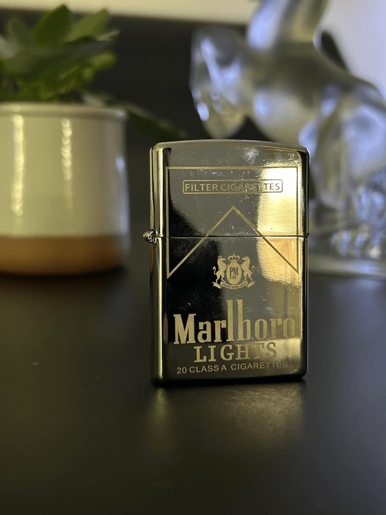 Puede incluir: Un encendedor plateado con el logotipo de Marlboro Lights. El encendedor tiene las palabras "FILTER CIGARETTES" en la parte superior y "Marlboro LIGHTS 20 CLASS A CIGARETTES" en la parte inferior. Est&aacute; sobre una superficie oscura.