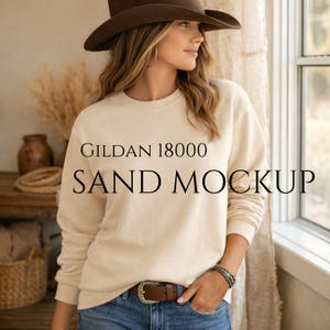 May include: A sand-colored Gildan 18000 sweatshirt mockup. The image features the text "GILDAN 18000 SAND MOCKUP" in black. The model is wearing a brown hat, blue jeans, and a brown belt.