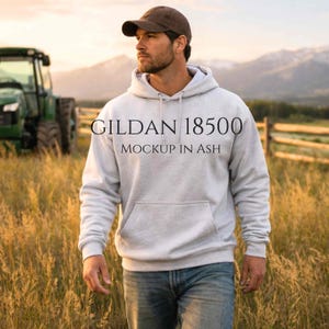Könnte beinhalten: Ein hellgrauer Kapuzenpullover mit dem Text "GILDAN 18500 MOCKUP IN ASH" wird von einer Person auf einem Feld getragen. Die Person trägt eine braune Baseballkappe und blaue Jeans. Ein grüner Traktor und ein Holzzaun sind im Hintergrund.