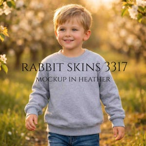 Può includere: Un bambino indossa una felpa grigio mélange e jeans blu. La felpa presenta la scritta "RABBIT SKINS 3317 MOCKUP IN HEATHER". Lo sfondo è una scena sfocata all'aperto con la luce del sole.