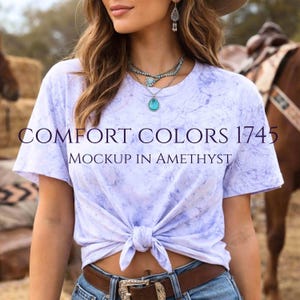 Puede incluir: Camiseta de color lila claro con nudo frontal y estampado veteado. La camiseta presenta el texto "COMFORT COLORS 1745 MOCKUP IN AMETHYST". La persona lleva un cinturón marrón con hebilla plateada y vaqueros.