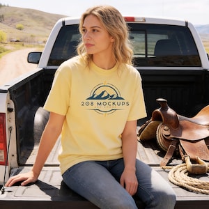 May include: A light yellow t-shirt with the text "208 Mockups and Goods" in a navy blue circular design. The shirt is short-sleeved and worn with blue jeans. A saddle and rope are visible in the background.