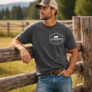May include: Dark gray t-shirt with a white logo featuring a cow silhouette and the text "208 MOCKUPS". The shirt is paired with blue jeans and a brown belt. The person is wearing a brown and tan baseball cap.