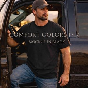 Puede incluir: Camiseta negra con el texto "COMFORT COLORS 1717 MOCKUP IN BLACK". La camiseta se usa con vaqueros azules y una gorra de béisbol marrón. La persona está de pie junto a un vehículo negro.