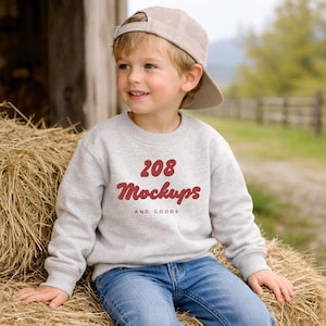 May include: A young child wearing a light gray sweatshirt and a tan baseball cap. The sweatshirt features the text "208 Mockups AND GOODS" in red lettering. The child is sitting on a bale of hay, wearing blue jeans.
