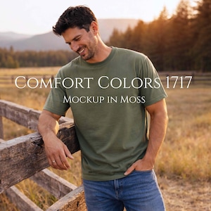 May include: A person wearing a moss green Comfort Colors 1717 t-shirt and blue jeans, leaning against a wooden fence. The text "COMFORT COLORS 1717 MOCKUP IN MOSS" is displayed on the shirt.