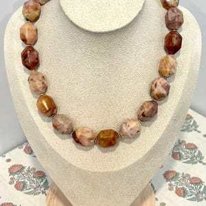 Op de afbeelding: Een ketting bestaande uit een enkele streng gefacetteerde, onregelmatige stenen kralen in tinten bruin, beige, crème en roodachtig oranje. De kralen zijn gepolijst en vertonen natuurlijke marmering en insluitingen in een cirkelvorm.