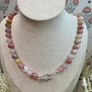 May include: A beaded necklace with a mix of pink, white, and yellow-toned beads. The necklace has a silver-colored clasp and a small white heart-shaped bead. The necklace is displayed on a mannequin.