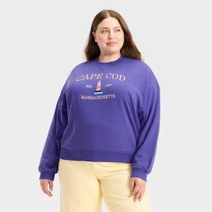May include: A purple sweatshirt featuring the text "CAPE COD" above a lighthouse graphic and "MASSACHUSETTS" below. The sweatshirt is worn with yellow trousers. The lighthouse graphic has red and white stripes.